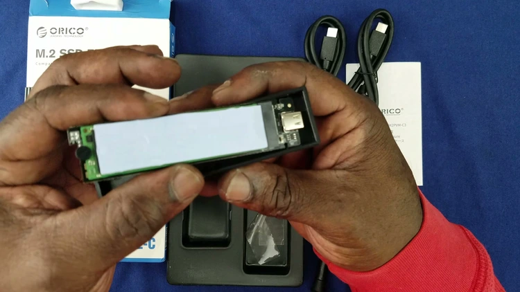 Placing the thermal pad over the NVMe SSD after removing the protective plastic film, with the drive seated in the open enclosure next to its USB-C port.